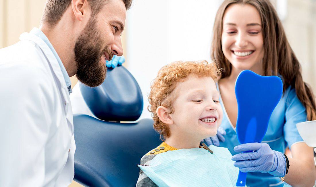 Smiling boy holding a mirror at the dentist with his mother and dentist